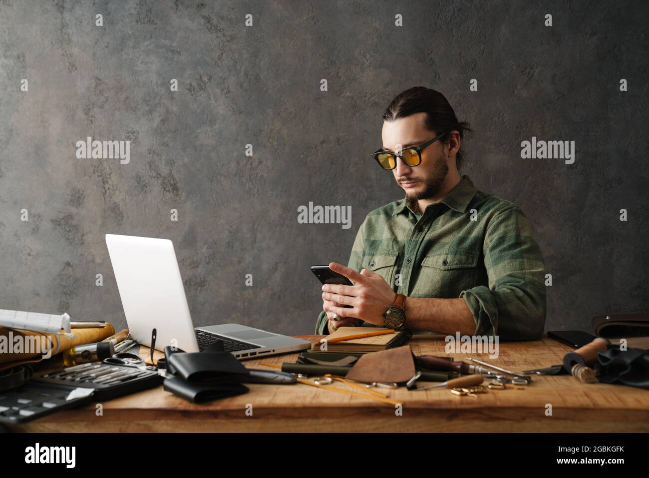 Bearded craftsman using laptop and smartphone while working with leather in workshop Stock Photo