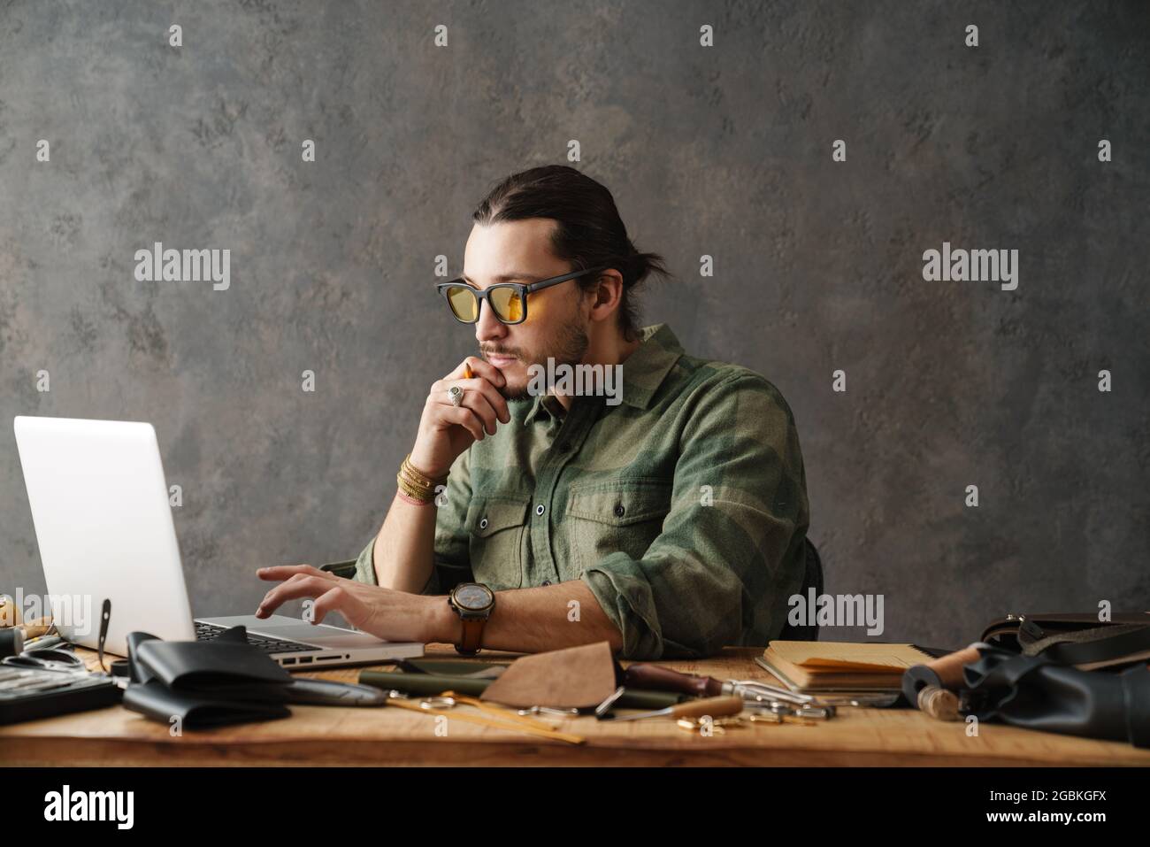 Bearded focused craftsman using laptop while working with leather in workshop Stock Photo