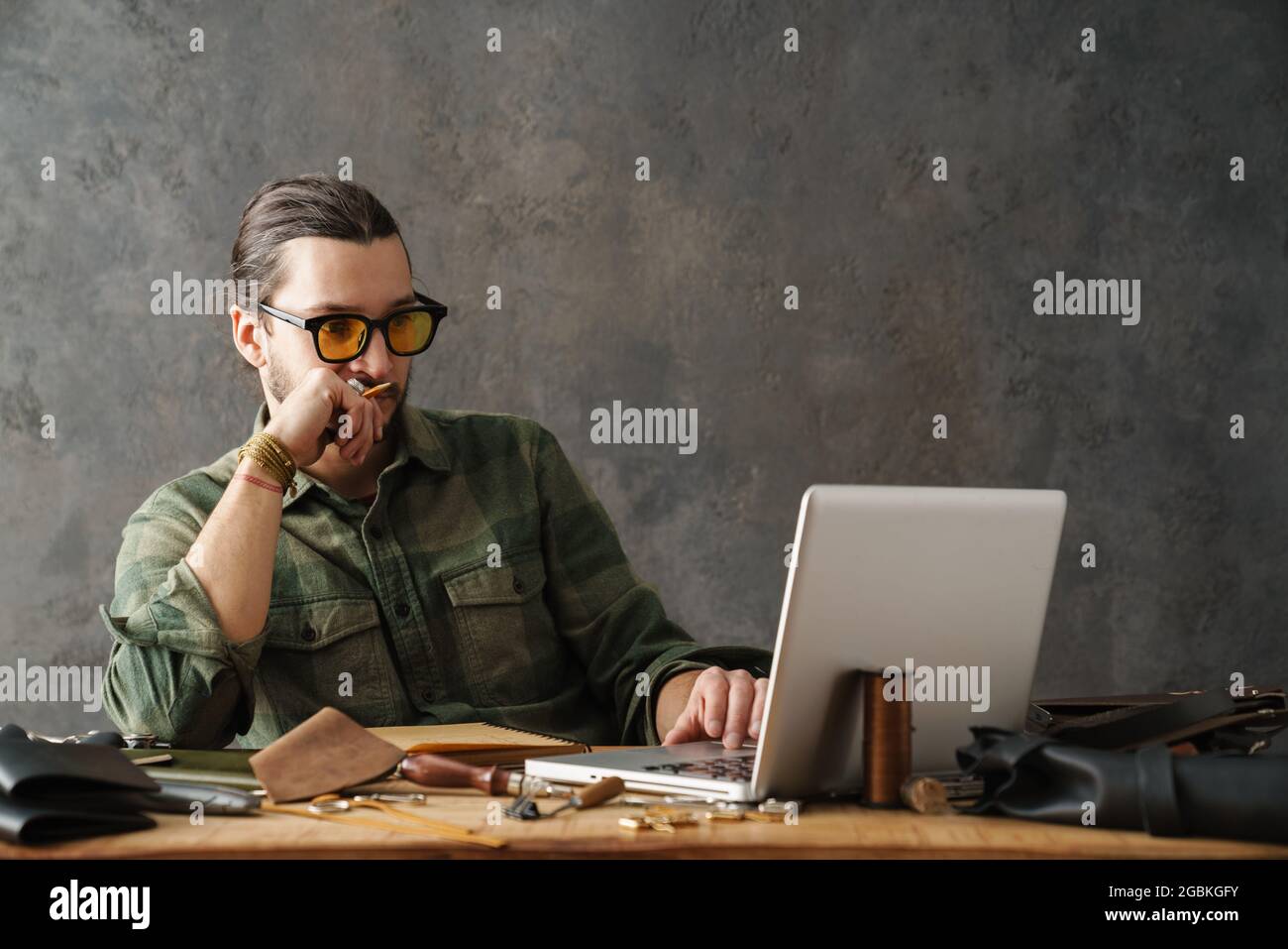Bearded focused craftsman using laptop while working with leather in workshop Stock Photo