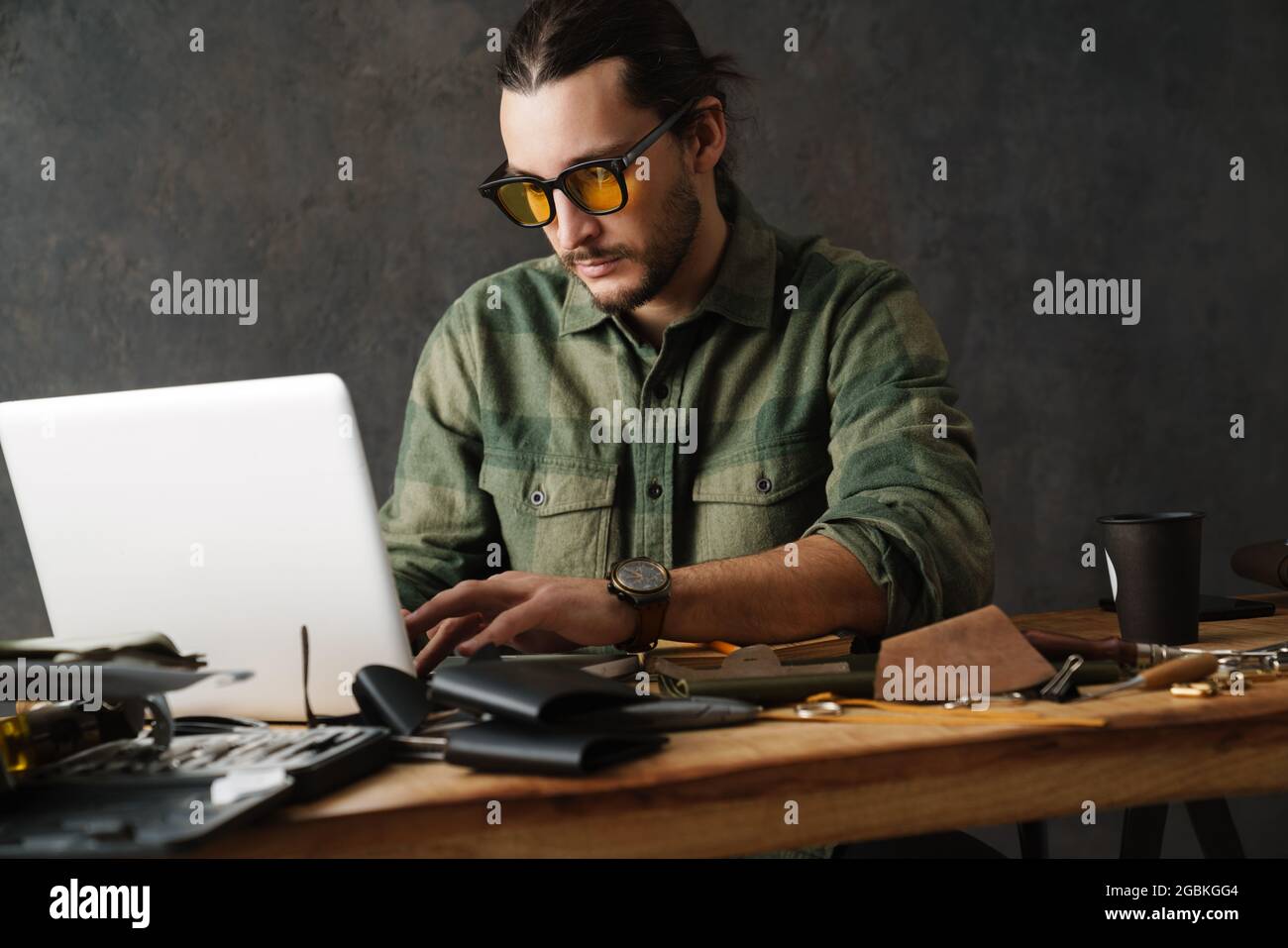 Bearded focused craftsman using laptop while working with leather in workshop Stock Photo
