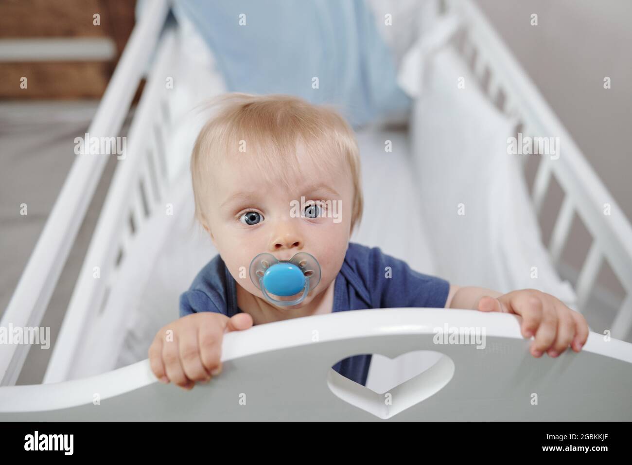 Portrait of serious cute blond-haired baby with dummy standing in cradle with heart-shaped hole Stock Photo