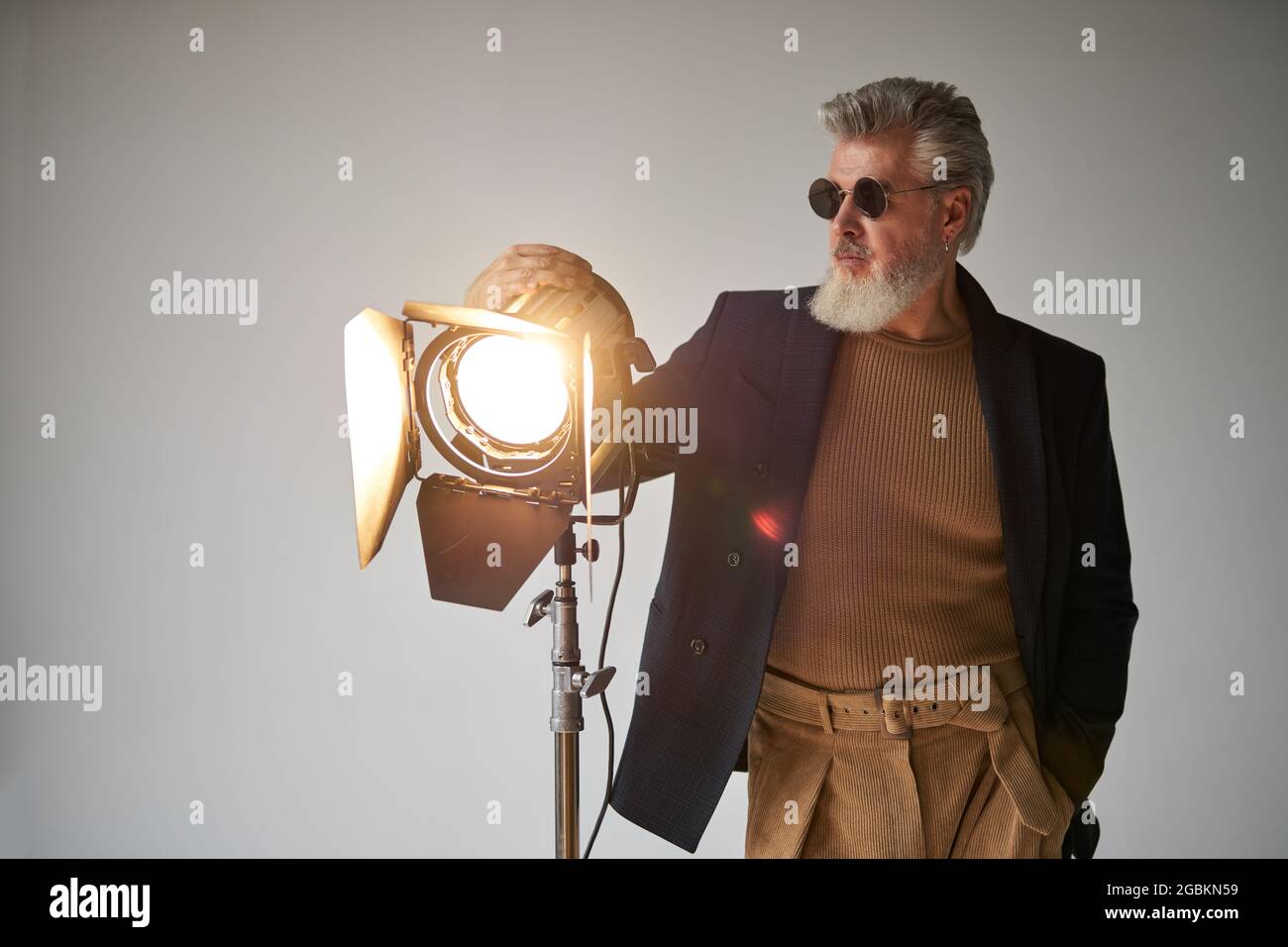 Portrait of trendy bearded middle aged man in glasses looking aside, standing next to studio spotlight while posing for camera over white background Stock Photo