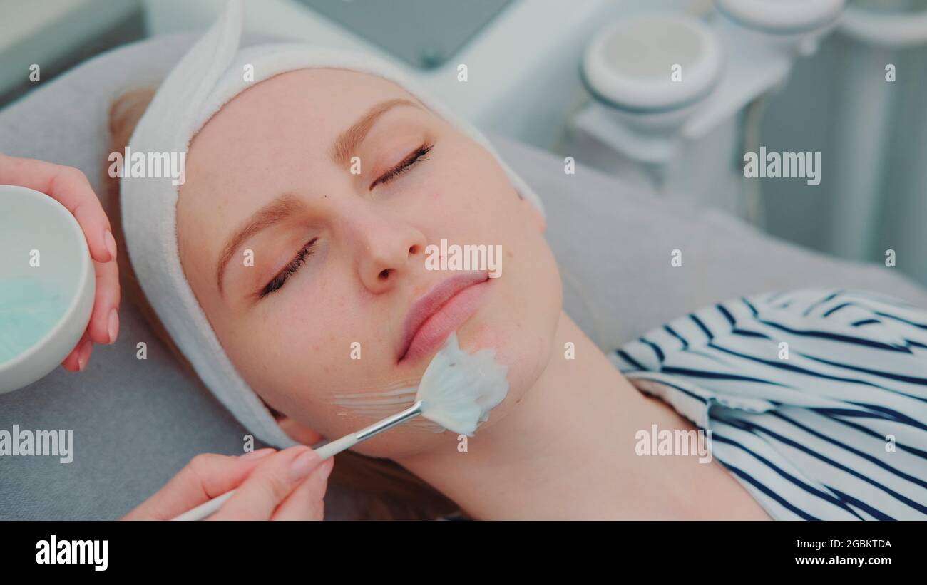 Cosmetician hands applying cream mask on young woman's face at beauty spa salon. Facial skin care treatments. Stock Photo