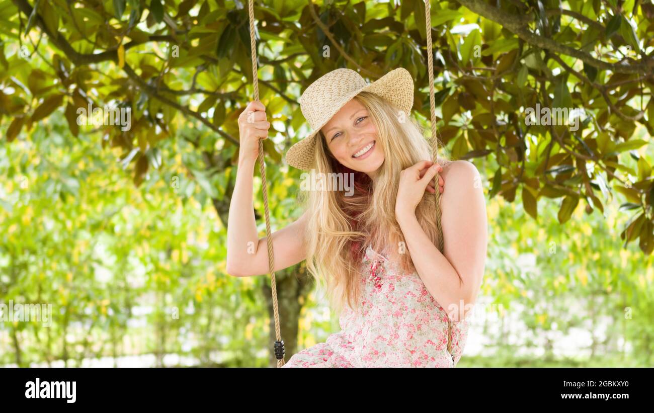 Smiling blonde woman wearing straw hat, swinging on the swing in the home garden, leisure and summer holiday concept Stock Photo