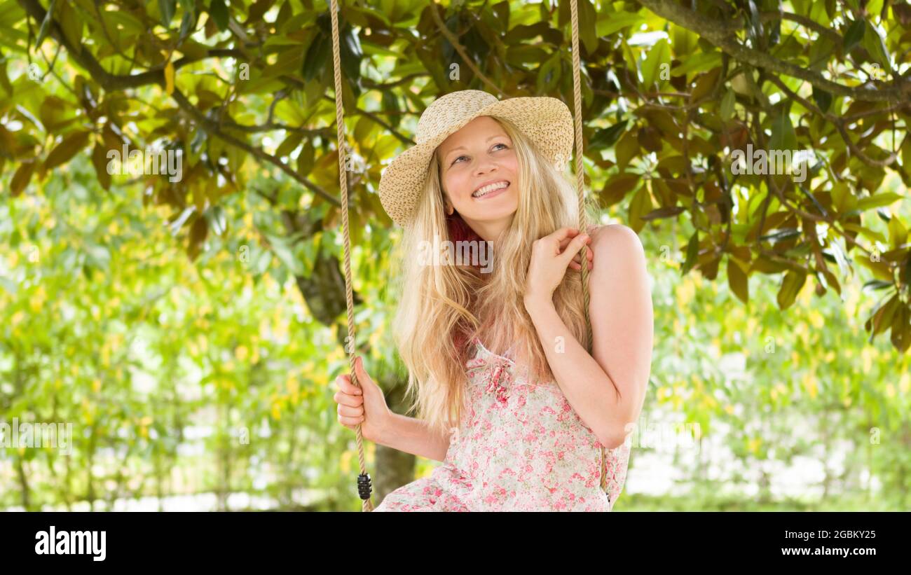 Smiling blonde woman wearing straw hat, swinging on the swing in the home garden, leisure and summer holiday concept Stock Photo