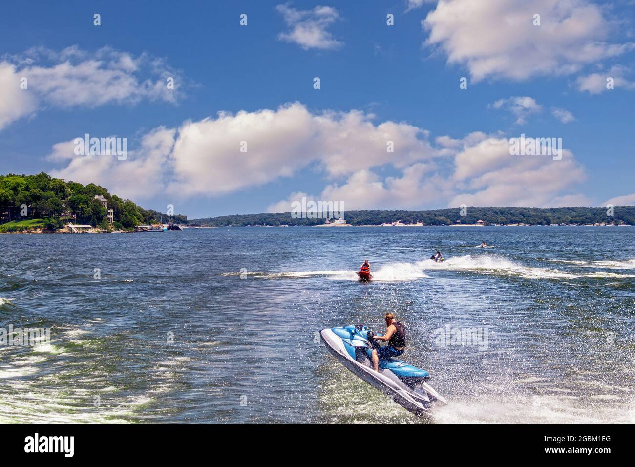 People on Personal watercraft jump wake of boat on lake with homes on shore in the distance. Stock Photo