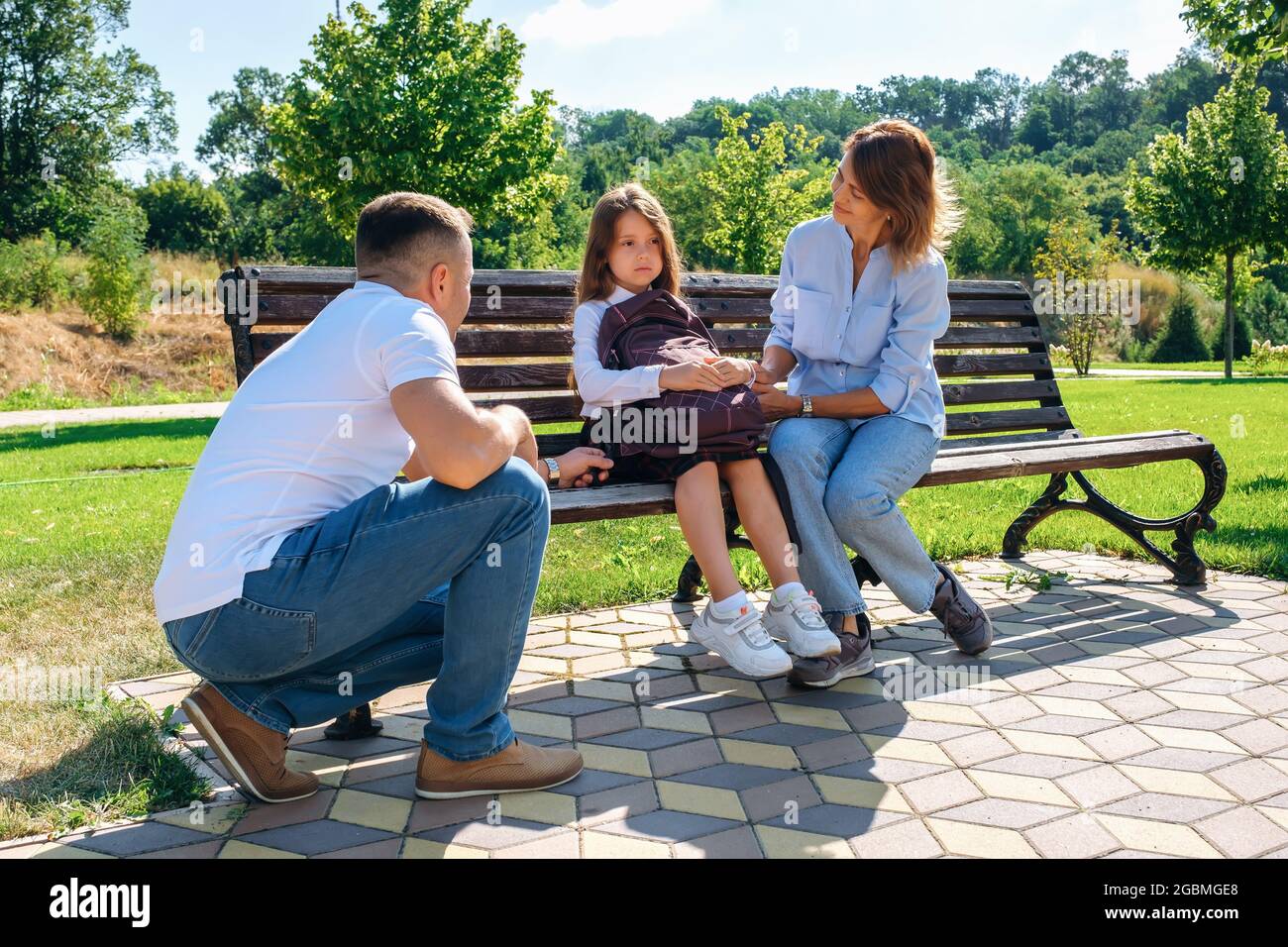 Unhappy schoolgirl sits with parents on a bench and does not want go to school Stock Photo