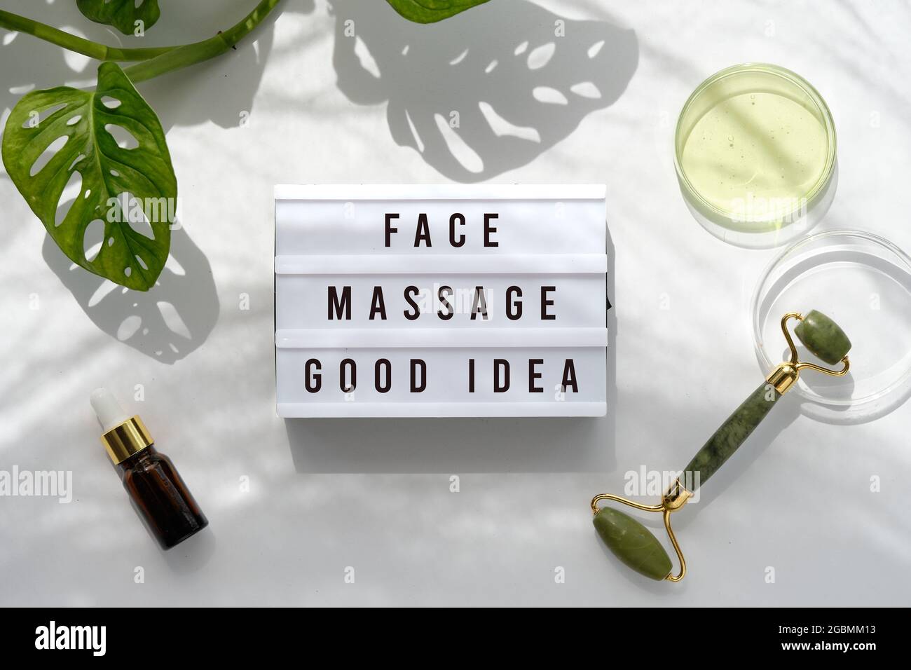Text Face Massage Good Idea on light box. Lightbox on Moisturizer, green jade face roller with exotic leaves. Monstera Adansonii leaves. Sunshine Stock Photo