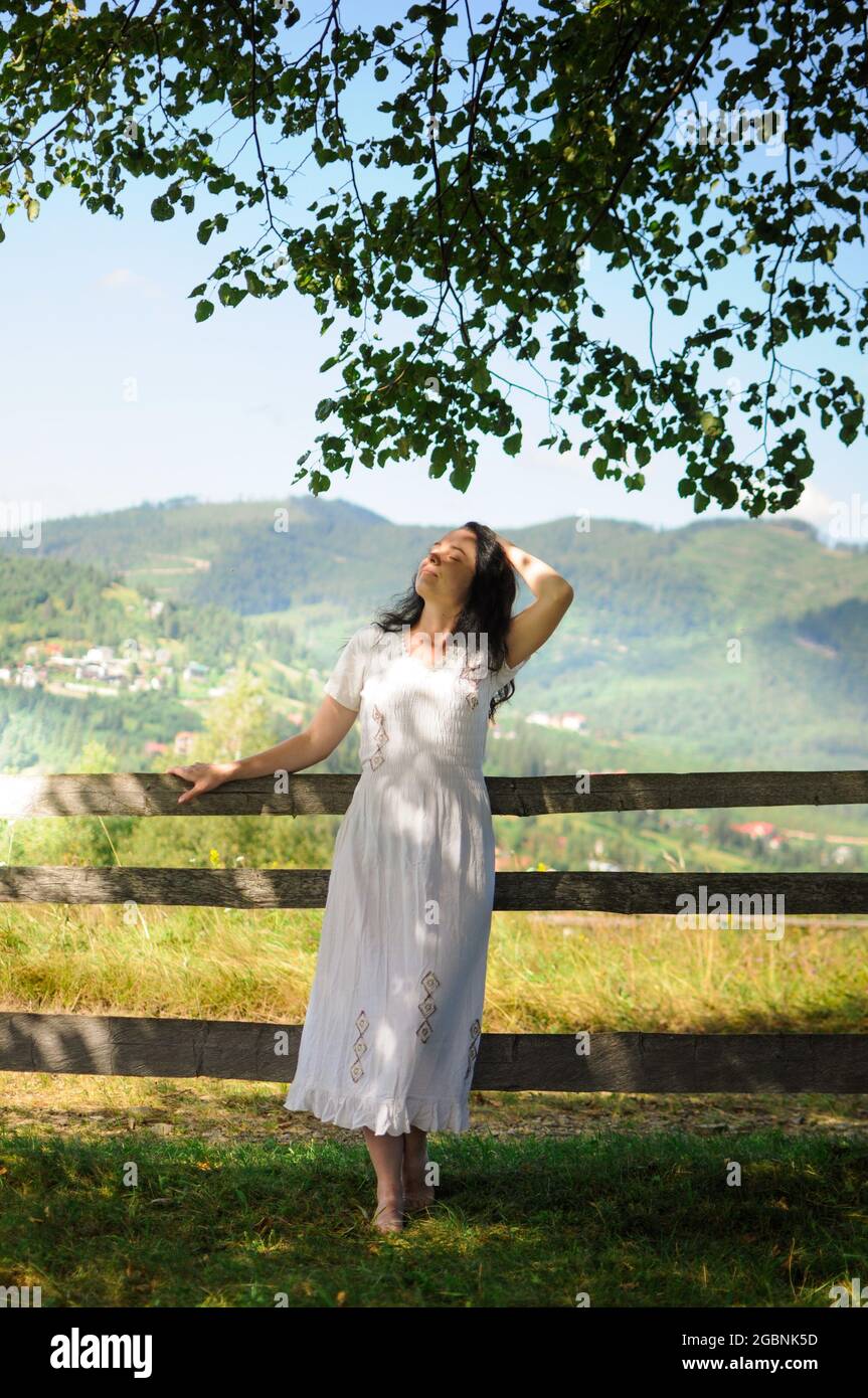 A young caucasian woman in a white ethnic dress is standing near a wooden fence on a mountain meadow and looking up. Natural style. Stock Photo