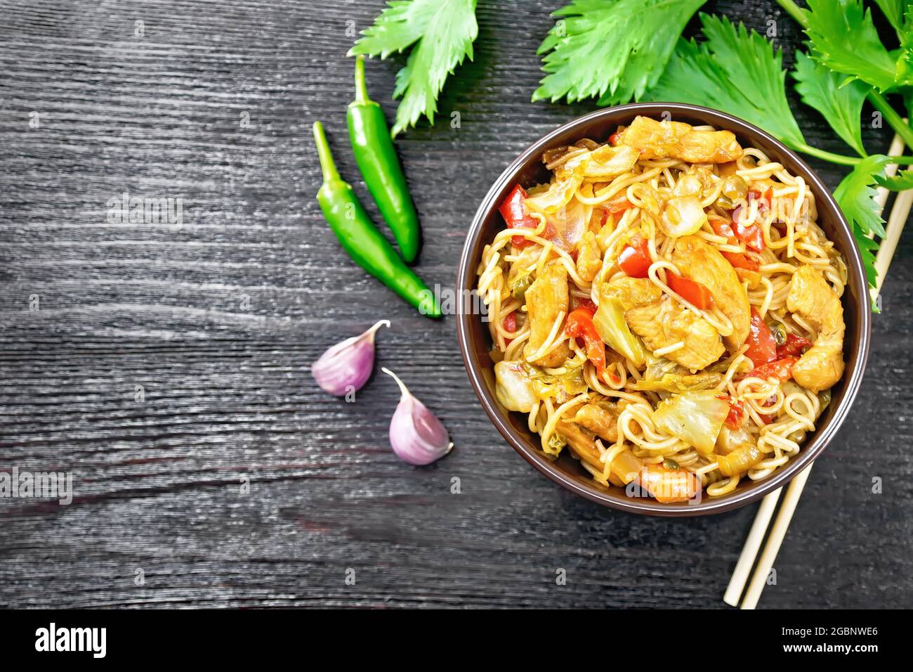 Noodles with cabbage, chicken breast, sweet red peppers, onions and peas seasoned with soy sauce in a bowl, garlic and celery leaves on dark wooden bo Stock Photo