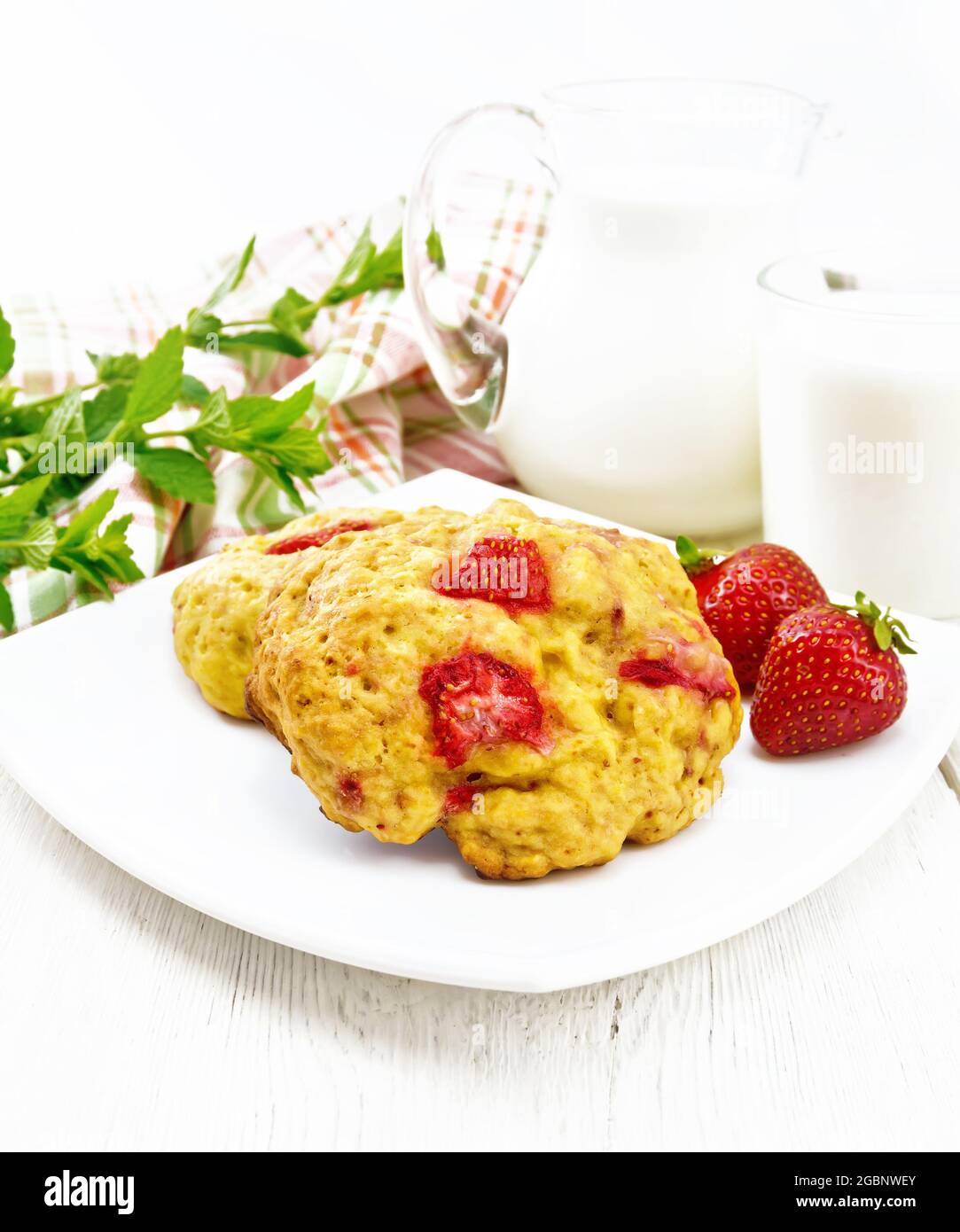 Strawberry scones in a plate with berries, a napkin, mint, milk in jug and glass on the background of light wooden board Stock Photo