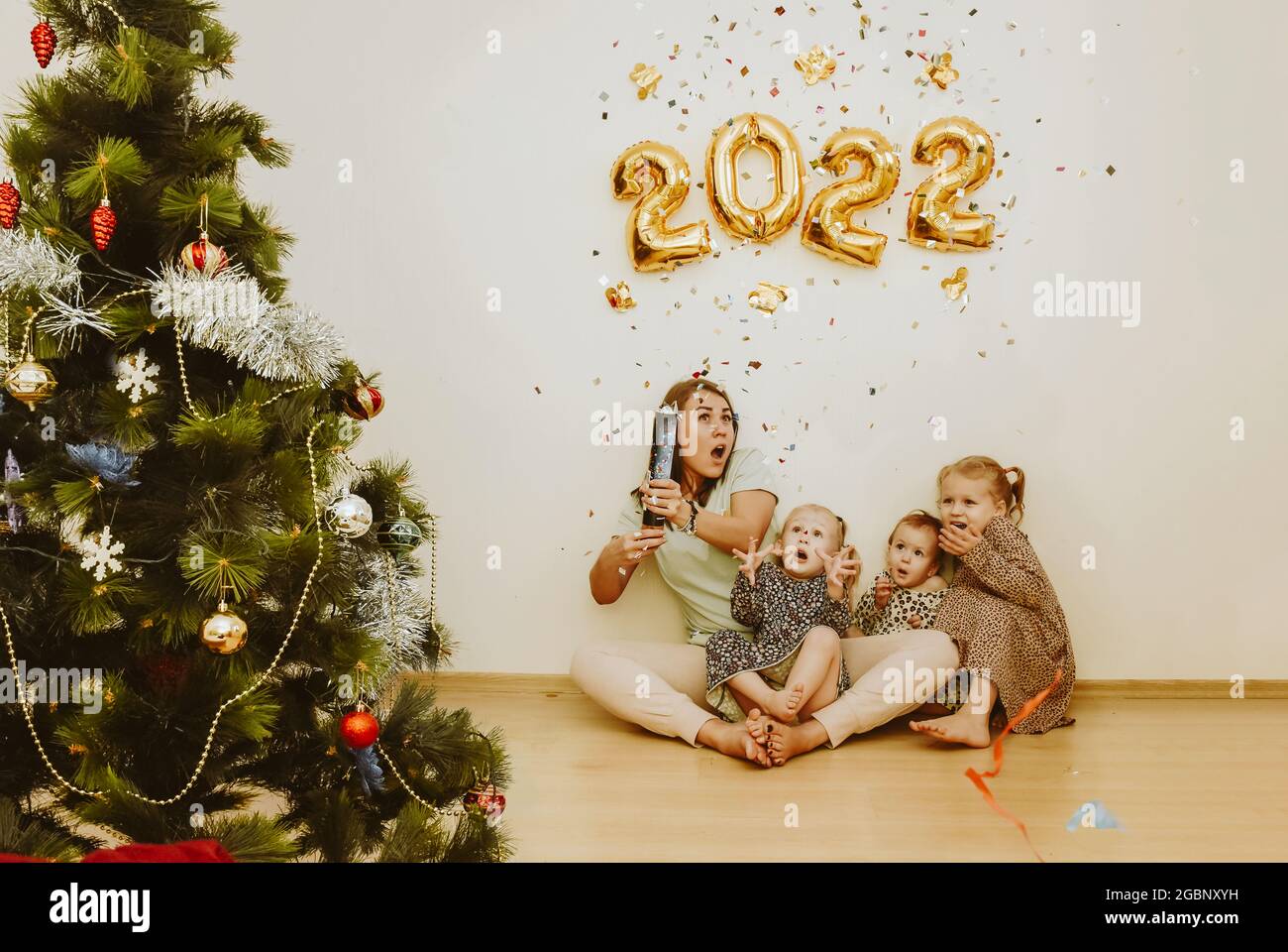 Mom and daughters with real emotions of joy explode a confetti clapper in the New Year 2022. Christmas family party at home with a Christmas tree and Stock Photo