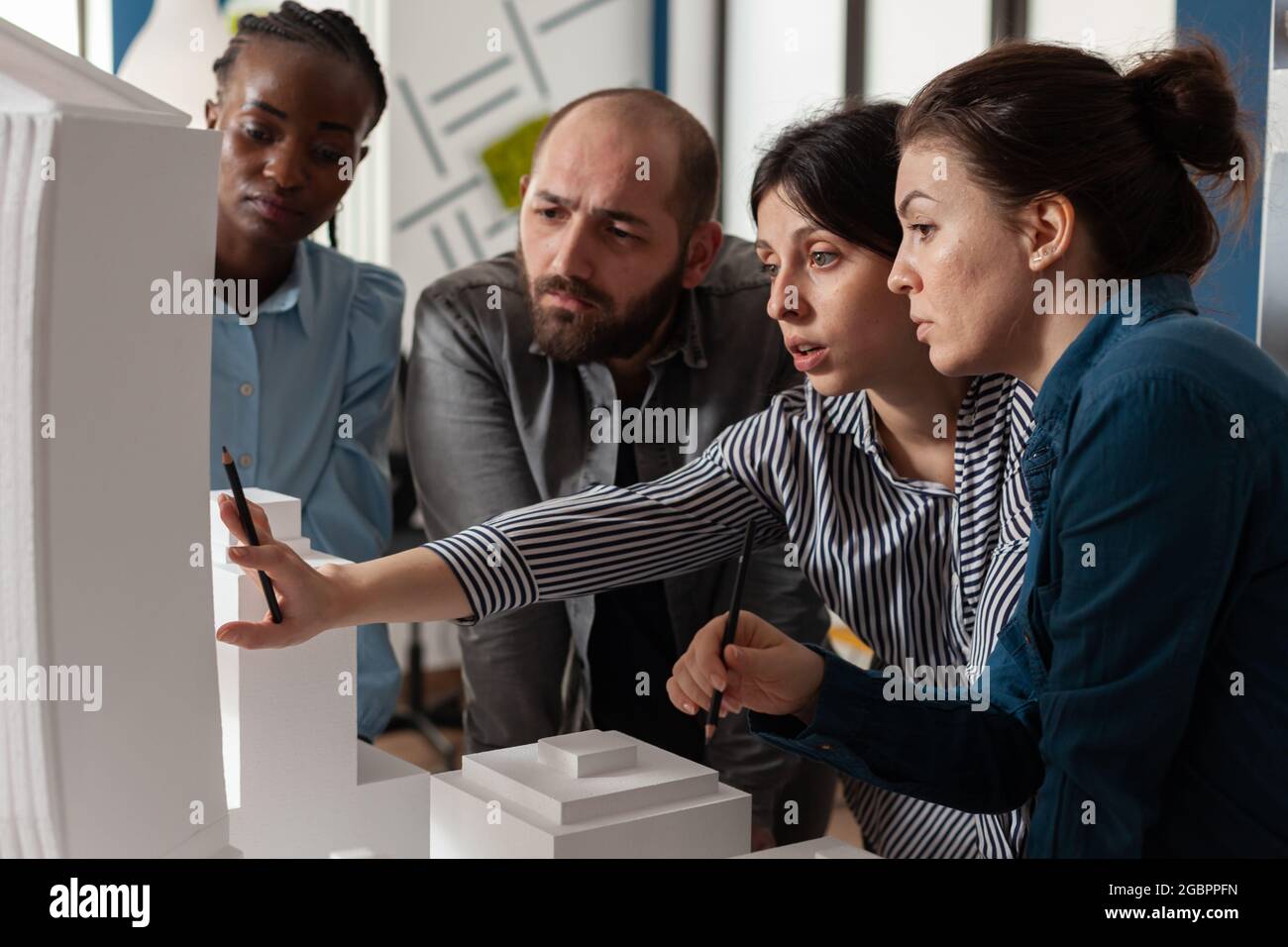 Multi ethnic architectural teamwork analyzing maquette building model construction layout. Diverse professional group of colleagues pointing at project design blueprint plan Stock Photo