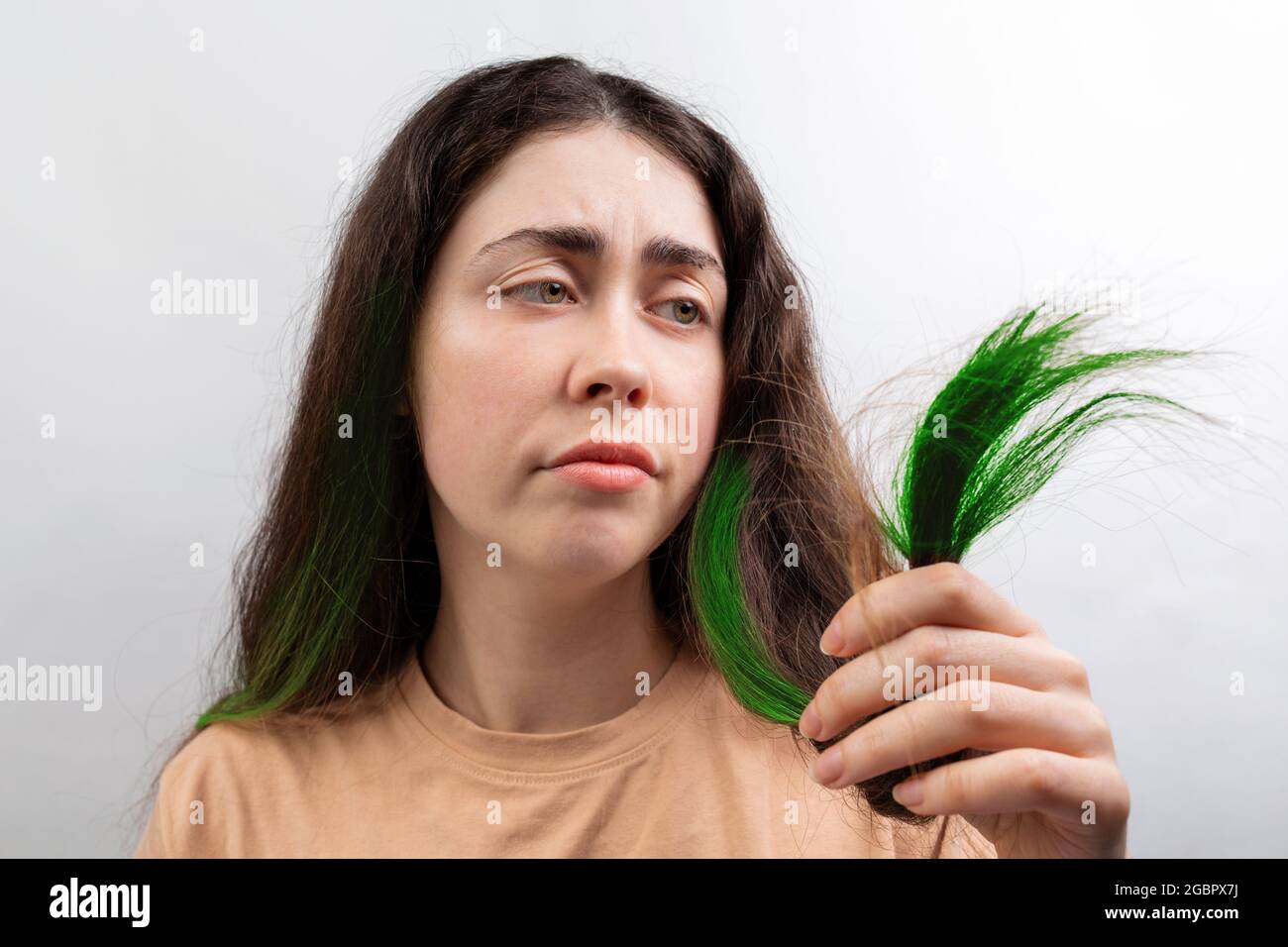 Hair coloring. A young Caucasian woman in a beige t-shirt looks distressed at the green tips of her long hair. White background. Stock Photo