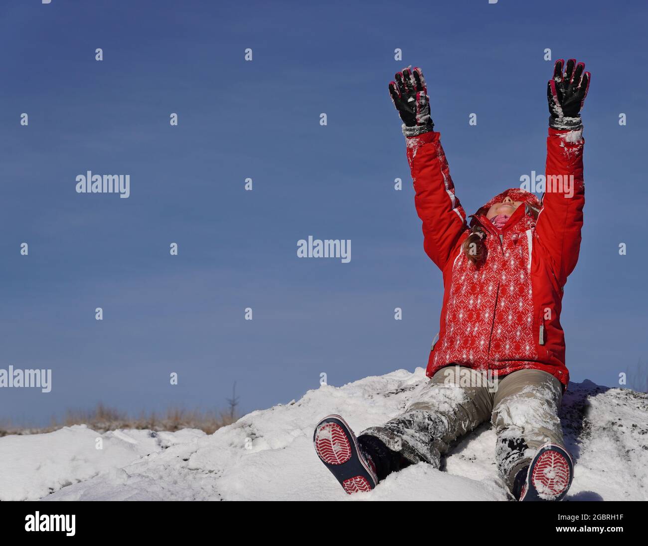 Happy kid sitting on the snow on sunny day rising arms up and looking to beautiful clear blue sky. Copyspace. Winter concept. Stock Photo