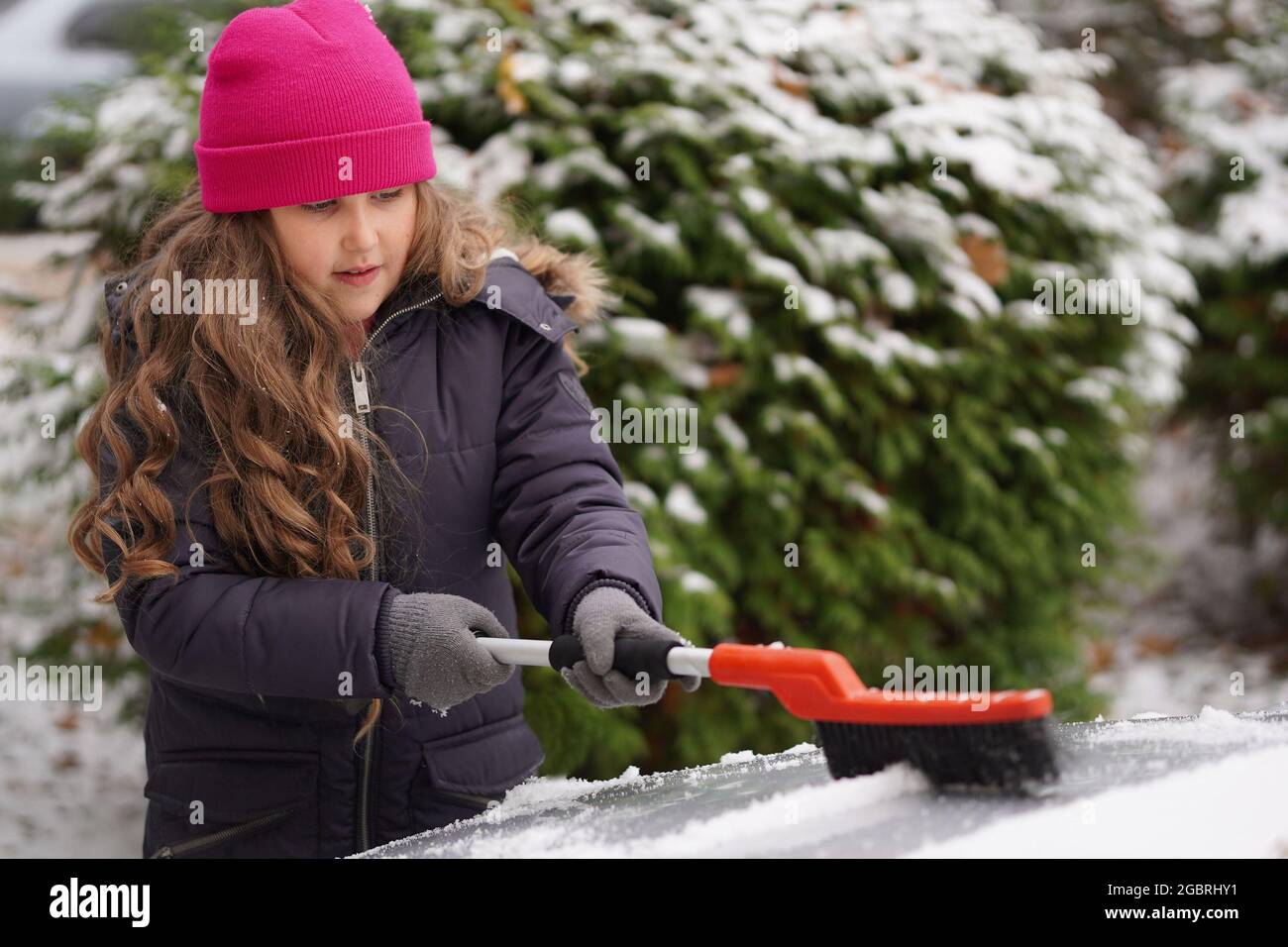 Adorable little curly girl helping to brush a snow from a car. Mommy's little helper. Winter activities for kids. Stock Photo