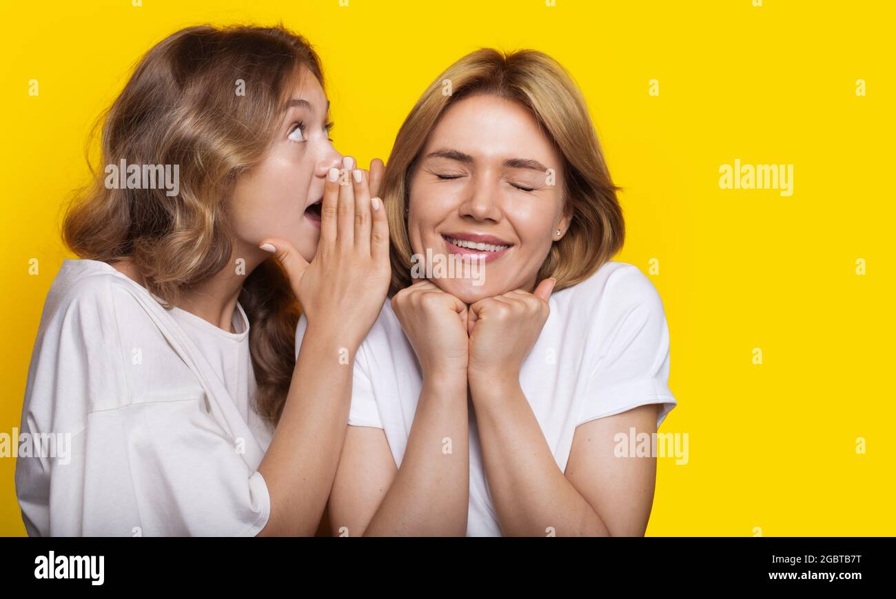 Blonde woman is whispering to her friend something while posing on a yellow studio wall Stock Photo