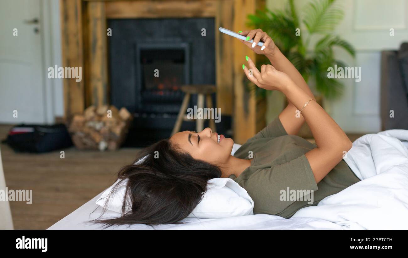 Smiling asian woman lies in bed with cell phone. She woke up and was ...