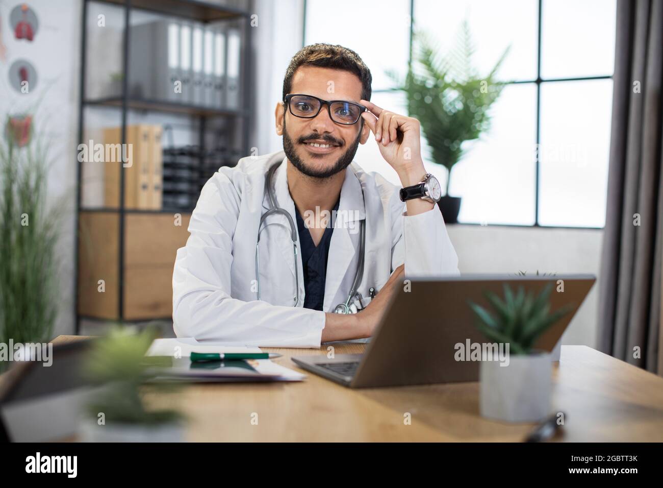 Close up of smiling handsome 30-aged male Malay doctor in eyeglasses, sitting at the table with laptop pc in his private office and looking at camera. Stock Photo