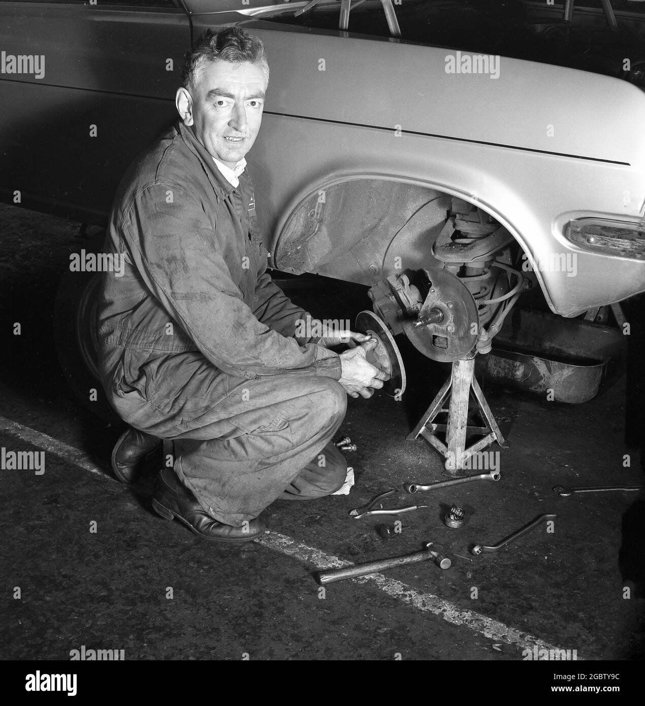 1975, historical, a car mechanic in overalls changing the disc brakes on a front wheel of a motorcar, which is propped up by a small jack or axle stand, England, UK. Stock Photo