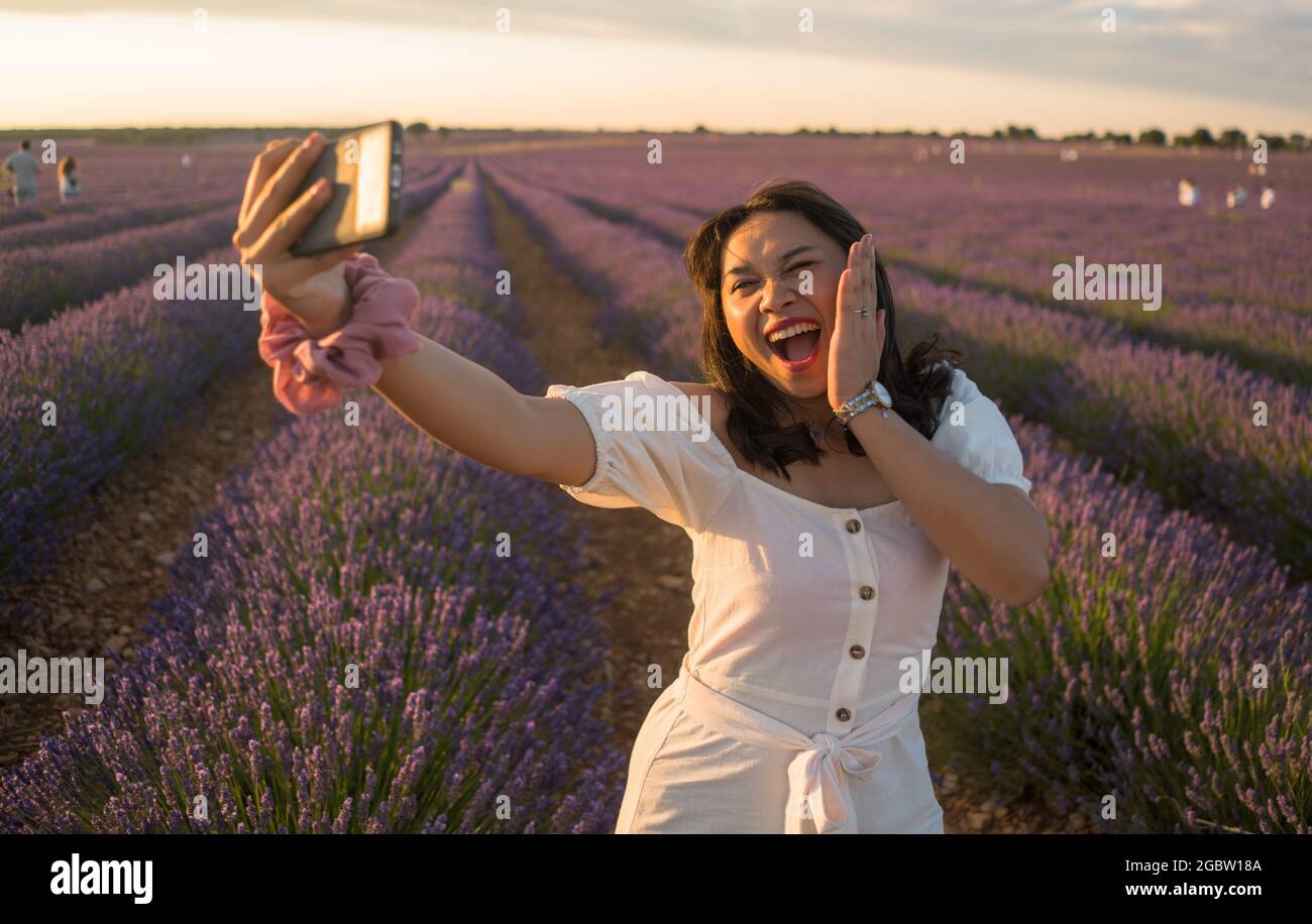 outdoors romantic portrait of young happy and attractive woman in white summer dress taking selfie with mobile phone at beautiful lavender flowers fie Stock Photo
