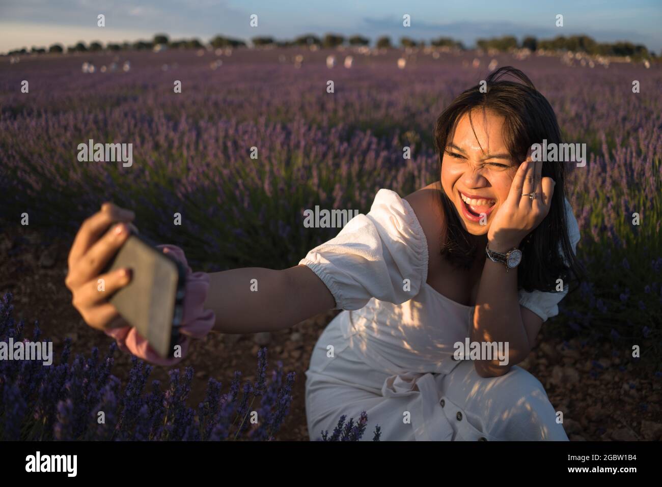 outdoors romantic portrait of young happy and attractive woman in white summer dress taking selfie with mobile phone at beautiful lavender flowers fie Stock Photo