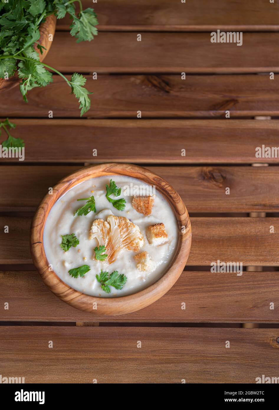Cream soup with white cauliflower, green herbs and croutons on top in wooden olive bowl on brown wooden table made of planks. Fresh cilantro on side. Stock Photo