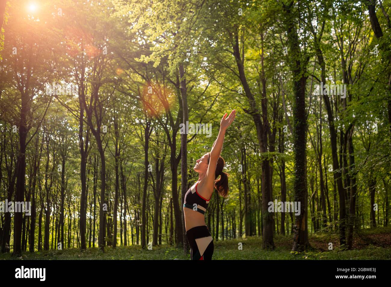 Sporty woman with her arms raised to the sun, forest setting, morning exercise. Stock Photo