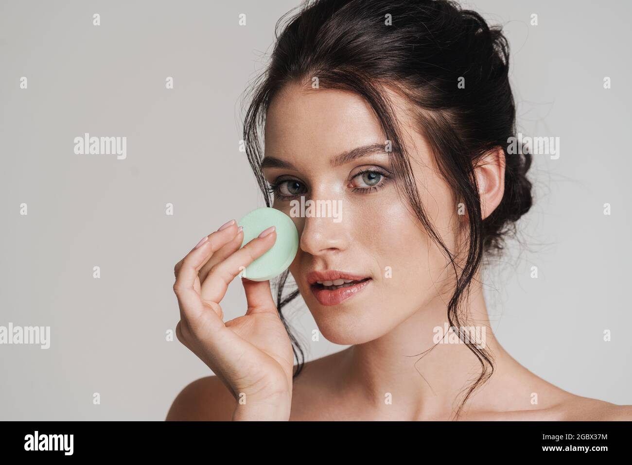 Young white woman with brown hair pulled up applying makeup using beauty blender sponge isolated over gray background Stock Photo