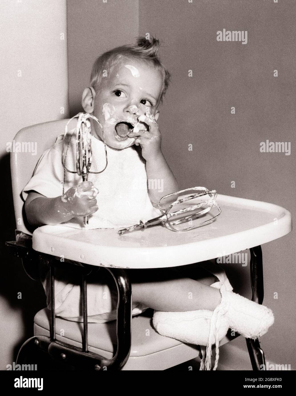 1950s BABY BOY WITH MESSY HANDS AND FACE SITTING IN HIGH CHAIR LICKING CAKE BATTER OFF OF ELECTRIC MIXER BEATERS  - b785 JHN001 HARS MALES ICING B&W BATTER GOALS HUMOROUS LICKING HIGHCHAIR MESSY COMICAL OPPORTUNITY CONCEPTUAL COMEDY BABY BOY BEATERS STICKY HIGH CHAIR JUVENILES BLACK AND WHITE CAUCASIAN ETHNICITY OLD FASHIONED Stock Photo