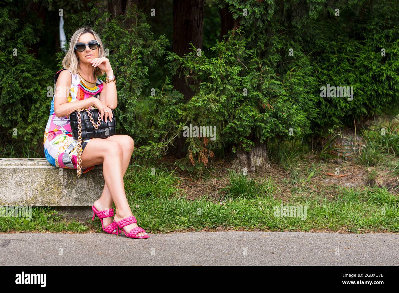 Blonde model sitting on stone bench in a colorful dress.The woman carries a black bag in her hand, sunglasses and pink sandals.The photograph was shot Stock Photo