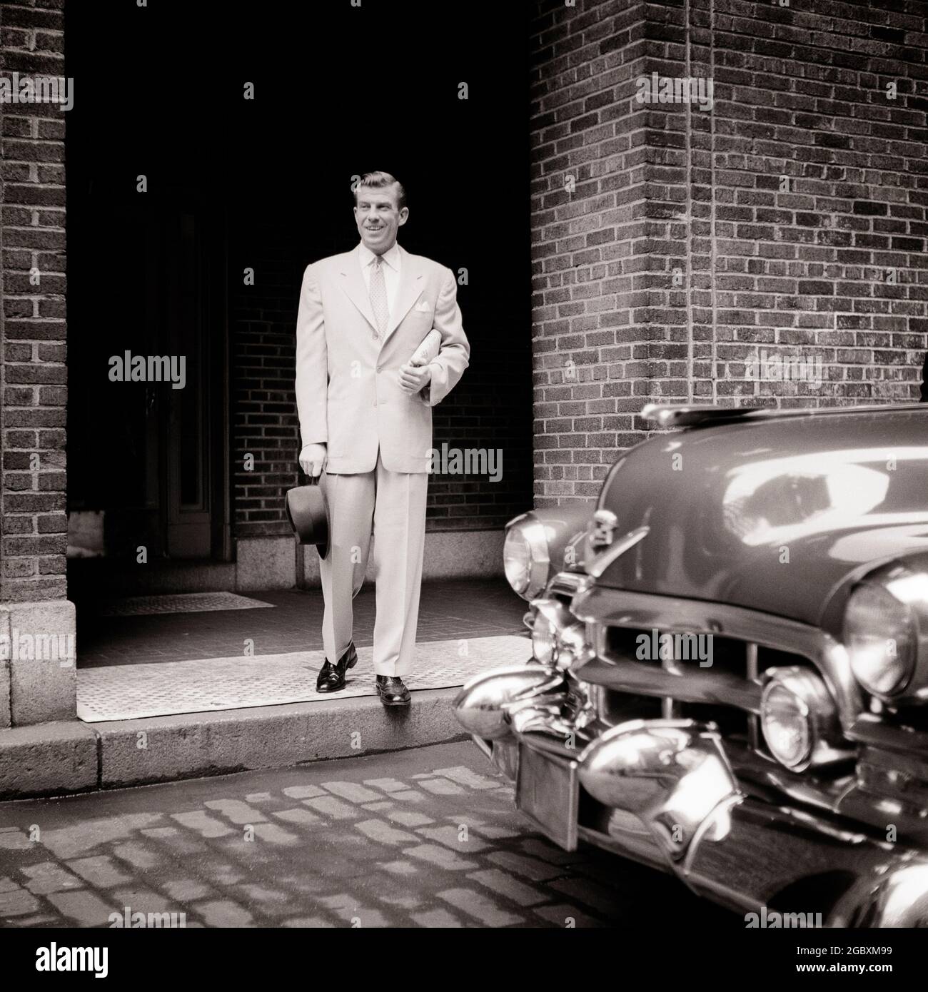 1950s DAPPER MAN DRESSED IN LIGHT SUIT TIE HAT STEPPING FROM BRICK BUILDING TO HIS CLASSIC CADILLAC AUTOMOBILE ON CITY STREET  - s6153 CRR001 HARS WEALTHY INFORMATION PLEASED JOY LIFESTYLE SATISFACTION BRICK STEPPING LUXURY COPY SPACE FULL-LENGTH PHYSICAL FITNESS PERSONS INSPIRATION AUTOMOBILE CARING MALES CONFIDENCE TRANSPORTATION B&W TEMPTATION SUIT AND TIE HAPPINESS CHEERFUL HIS LEISURE STRENGTH AUTOS CHOICE EXTERIOR LEADERSHIP LOW ANGLE POWERFUL PROGRESS PRIDE AUTHORITY OCCUPATIONS SMILES CONCEPTUAL AUTOMOBILES CADILLAC DAPPER JOYFUL VEHICLES WELL TO DO CREATIVITY IDEAS MID-ADULT Stock Photo