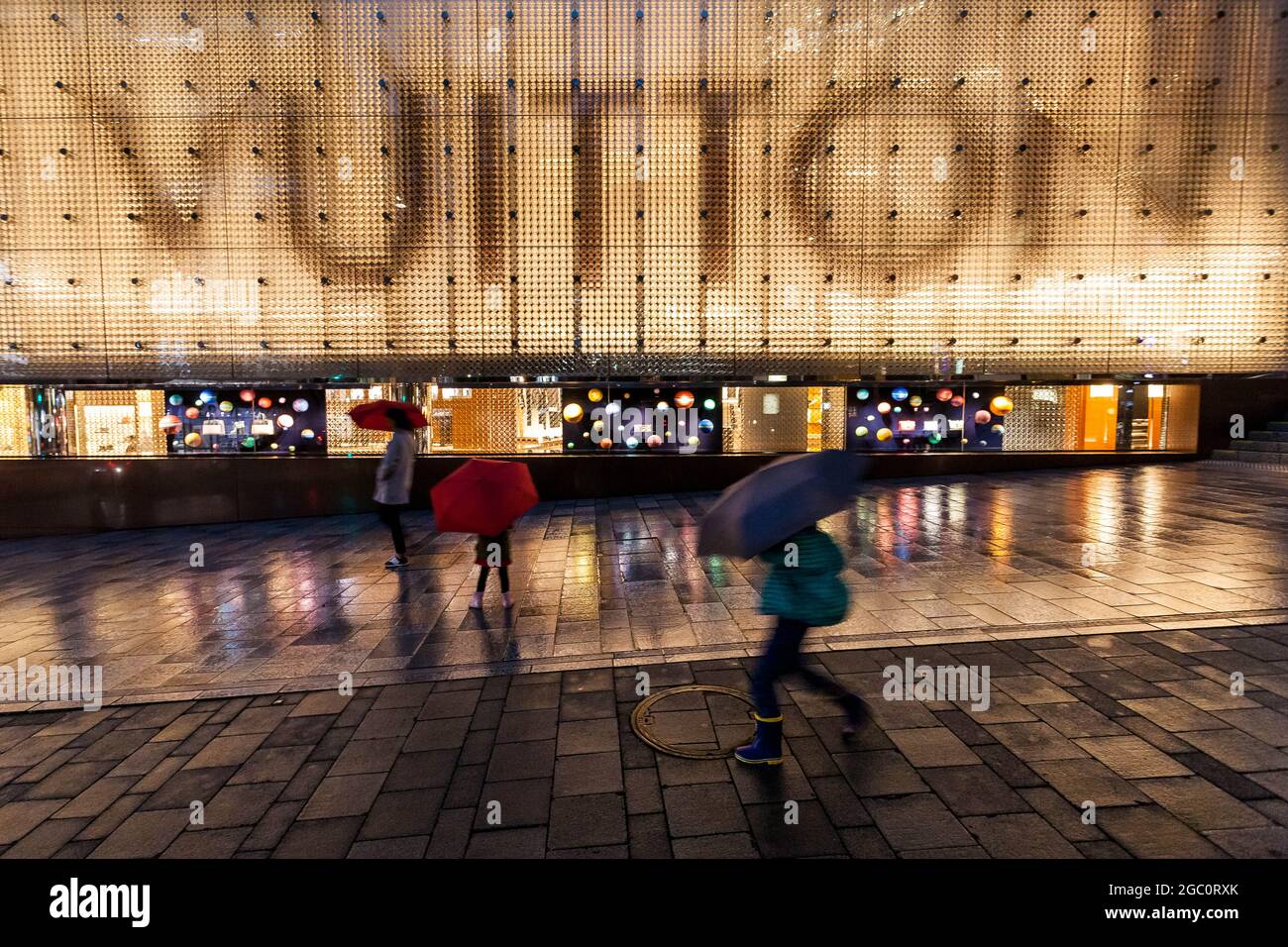 People walk past Louis Vuitton store in rain at night in Tokyo