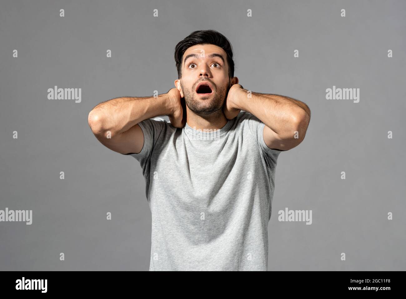 Shocked young Caucasian man gasping with hands around neck looking upward on light gray studio background Stock Photo