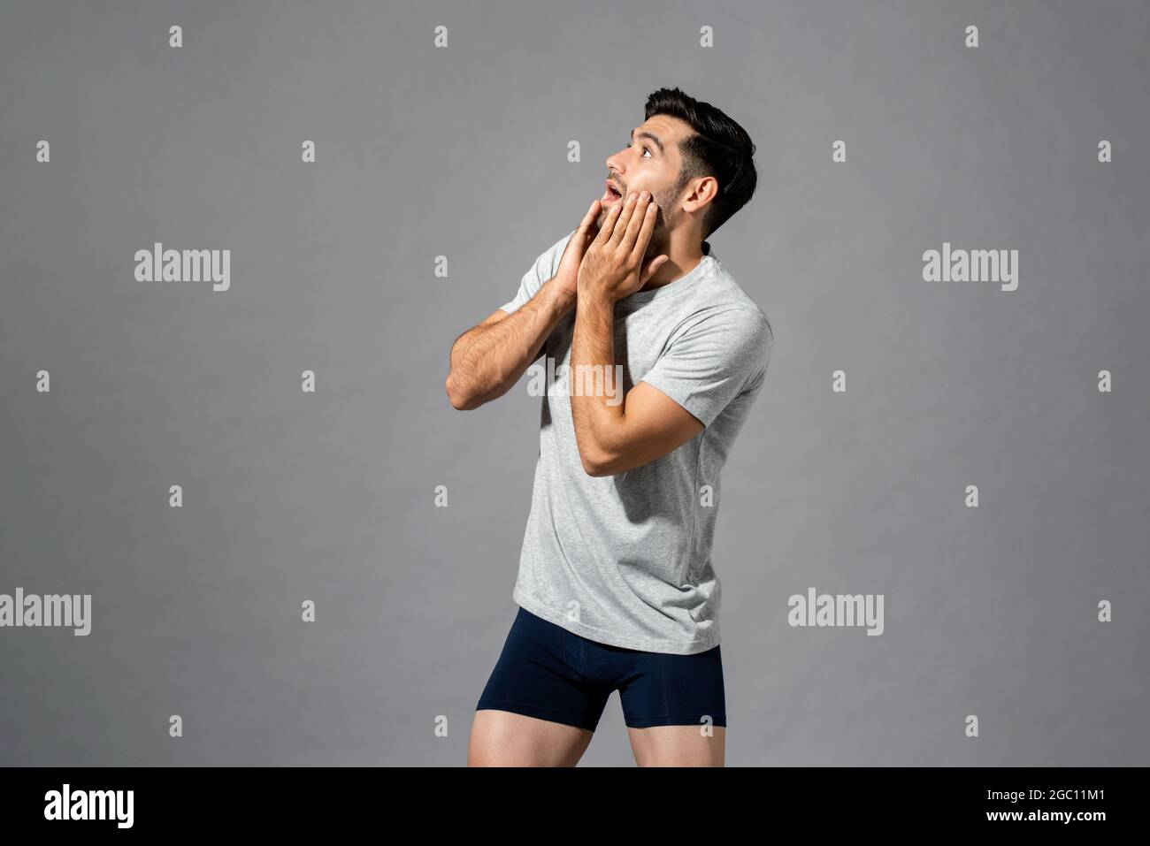 Surprised young Caucasian man in sleepwear with hands on cheeks looking up to empty space aside in light gray isolated studio background Stock Photo