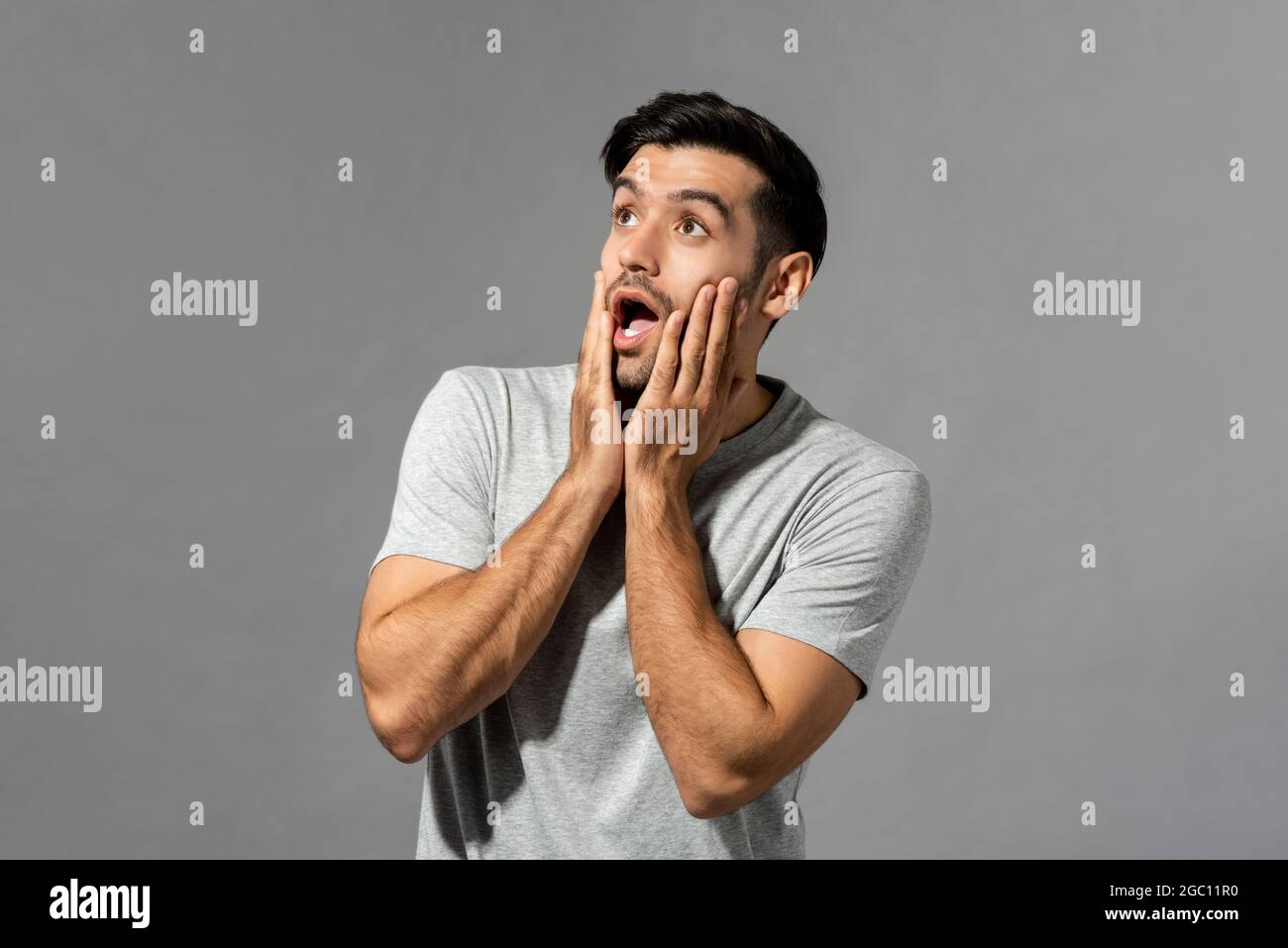 Shocked young Caucasian man with hands on cheeks gasping and looking up in light gray isolated studio background Stock Photo