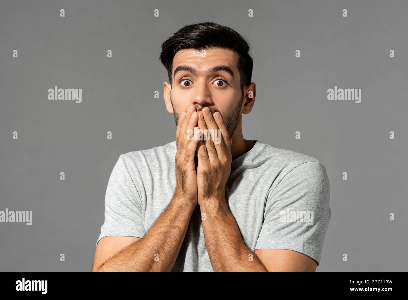 Shocked scared young Caucasian man with eyes popping and hands covering mouth on light gray studio background Stock Photo