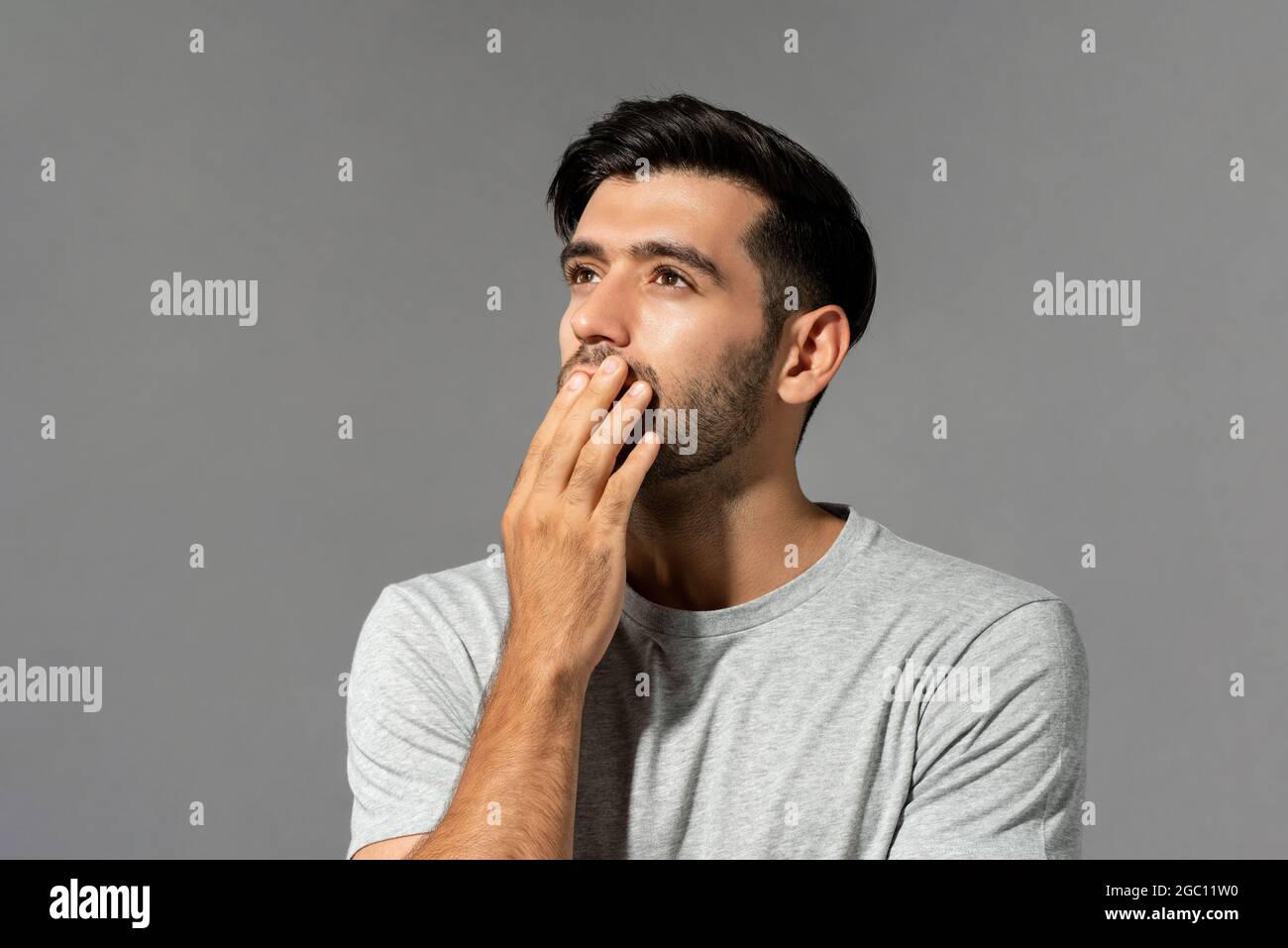 Shocked young Caucasian man looking up and thinking with hand cover mouth on light gray studio background Stock Photo