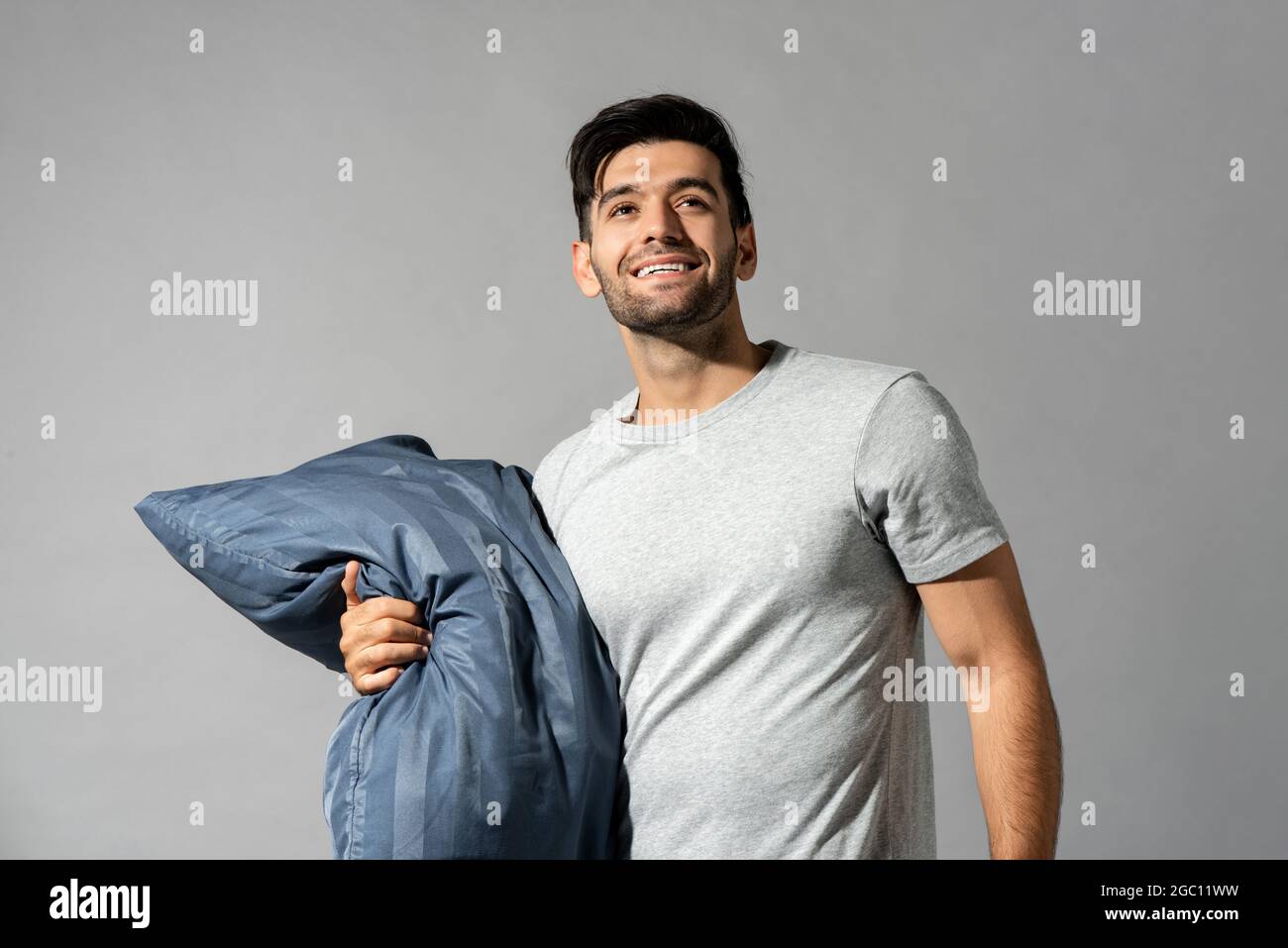 Smiling fresh young man in sleepwear holding pillow looking upward in isolated light gray studio background Stock Photo