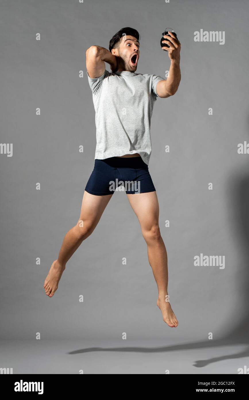 Full length portrait of shocked young Caucasian man in sleepwear waking up late looking at alarm clock and jumping in isolated gray studio background Stock Photo