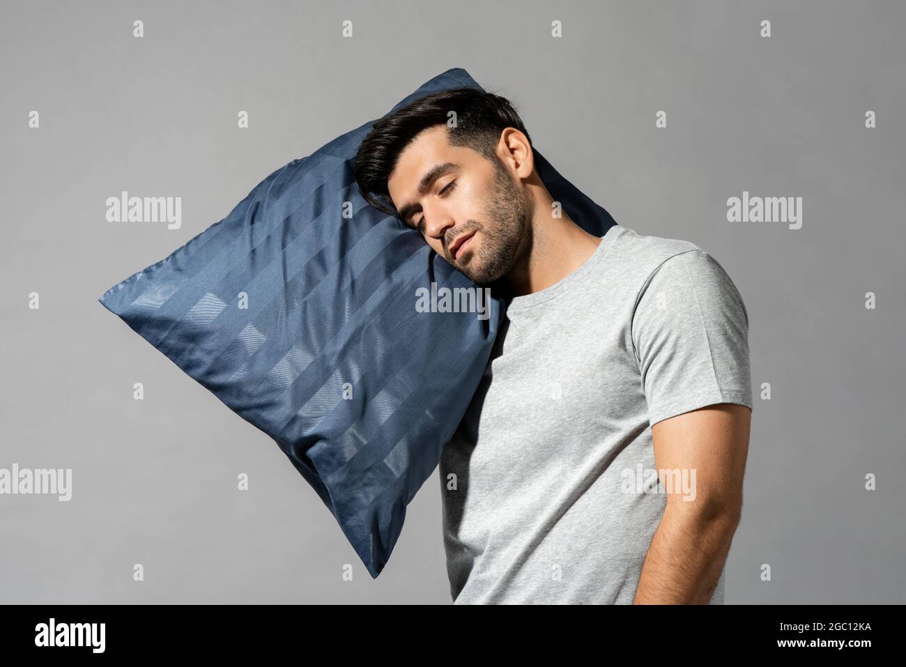 Young sleeping man holding his pillow standing in light gray isolated studio background, sleepwalking concept Stock Photo