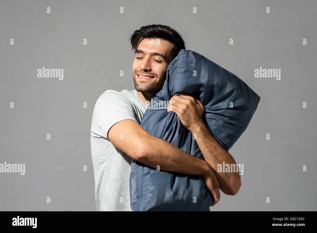 Sleepy young man holding pillow closing his eyes dreaming and smiling in isolated light gray studio background Stock Photo