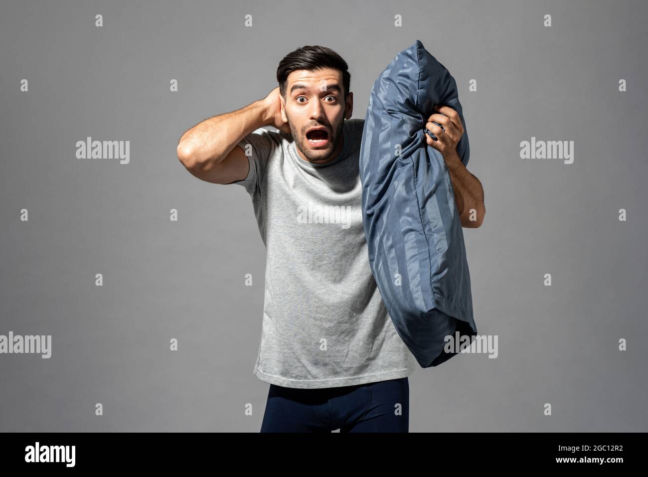 Young Caucasian man in sleepwear holding pillow waking up late and being shocked in light gray isolated studio background Stock Photo