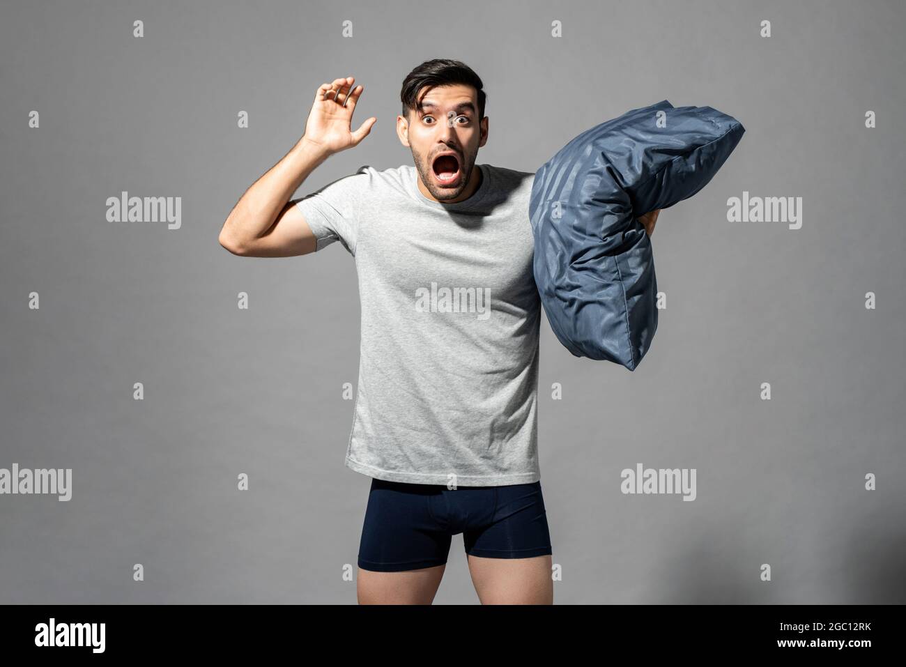 Sleepy young man in sleepwear feeling scared by nightmare and holding pillow, studio shot in isolated gray background Stock Photo