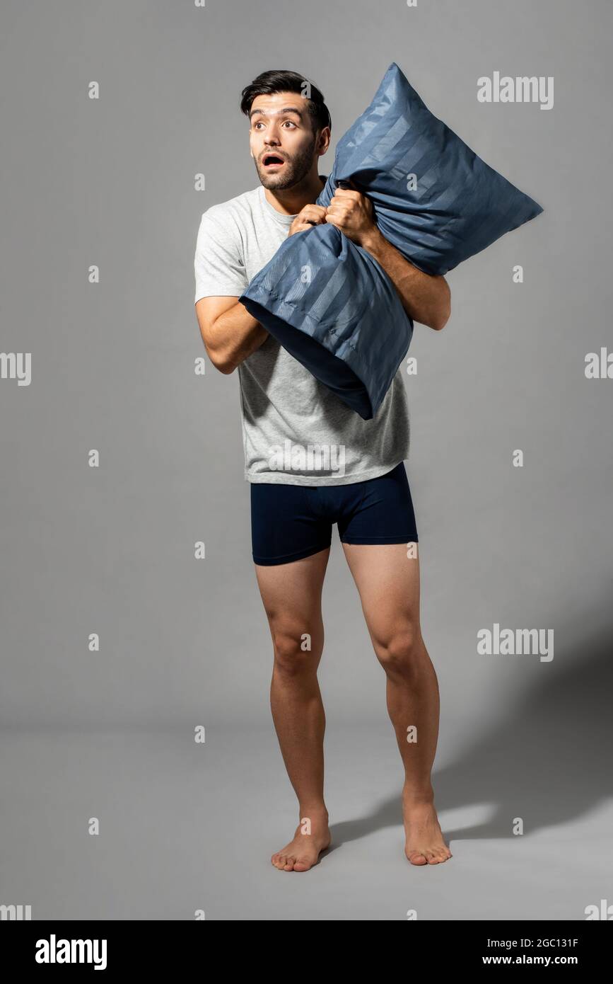 Full lenght portrait of young Caucasian man holding pillow and being shocked after waking up, studio shot in light gray isolated background Stock Photo