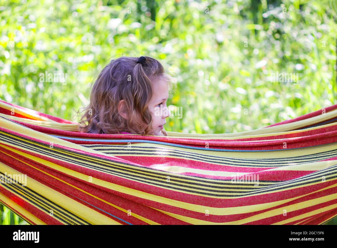 Adorable little girl with curly hair in colorful striped hammock on summer nature background in countryside. Stock Photo