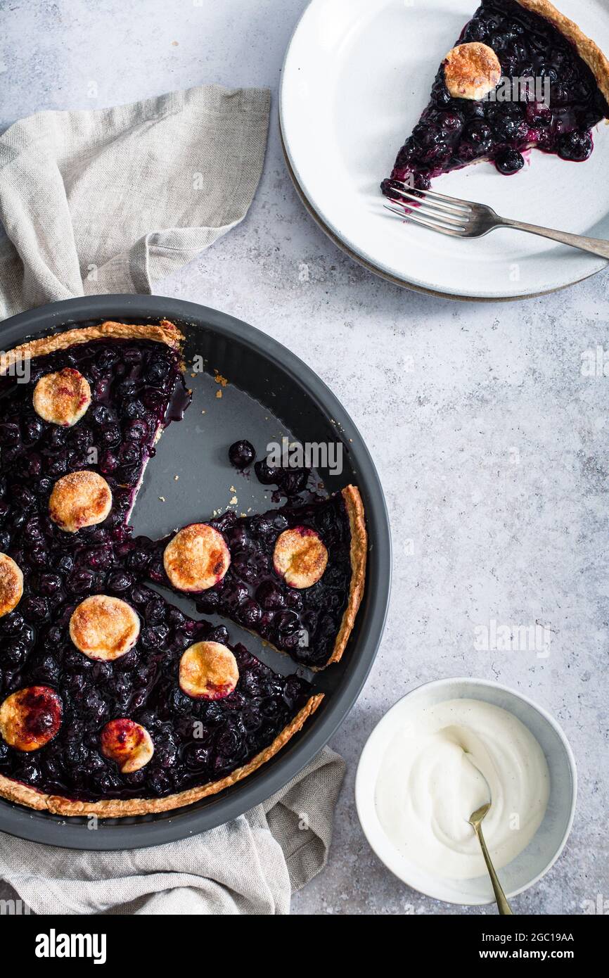 Sliced blueberry tart in its tin with a slice on a plate with a fork and a small bowl of whipped cream. Stock Photo