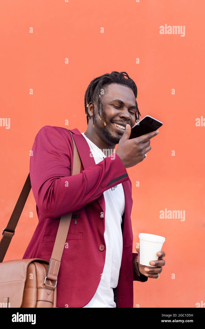 smiling african business man dressed in suit with briefcase and coffee records a voice message on his phone in front of a red background, copy space f Stock Photo
