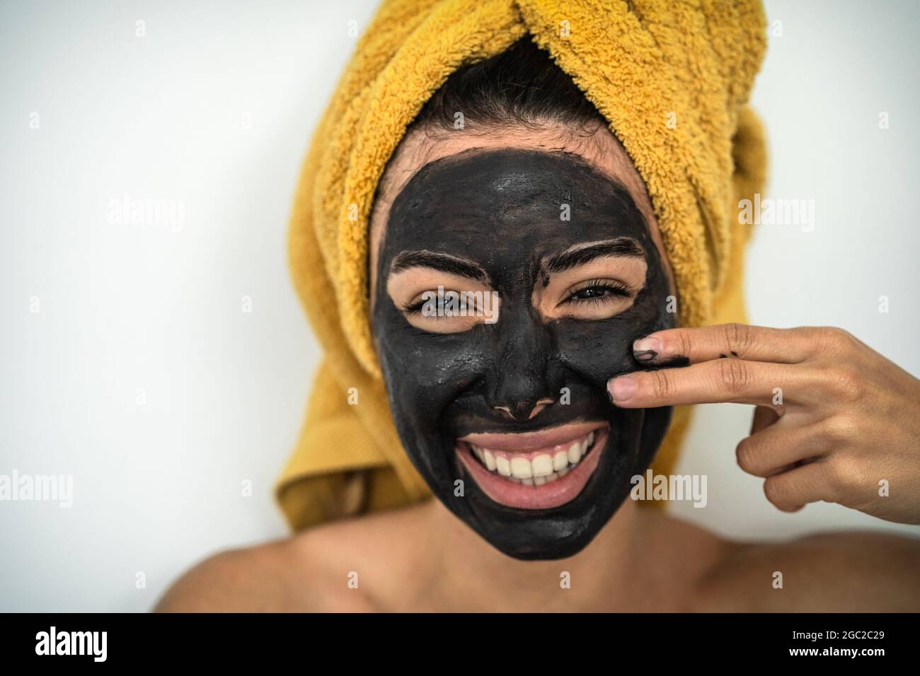 Young smiling woman applying charcoal mask on face - Happy girl having skin care spa day at home Stock Photo