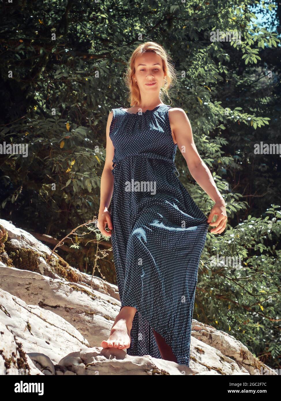 A girl in long dress walks barefoot along a rocky area under green ...